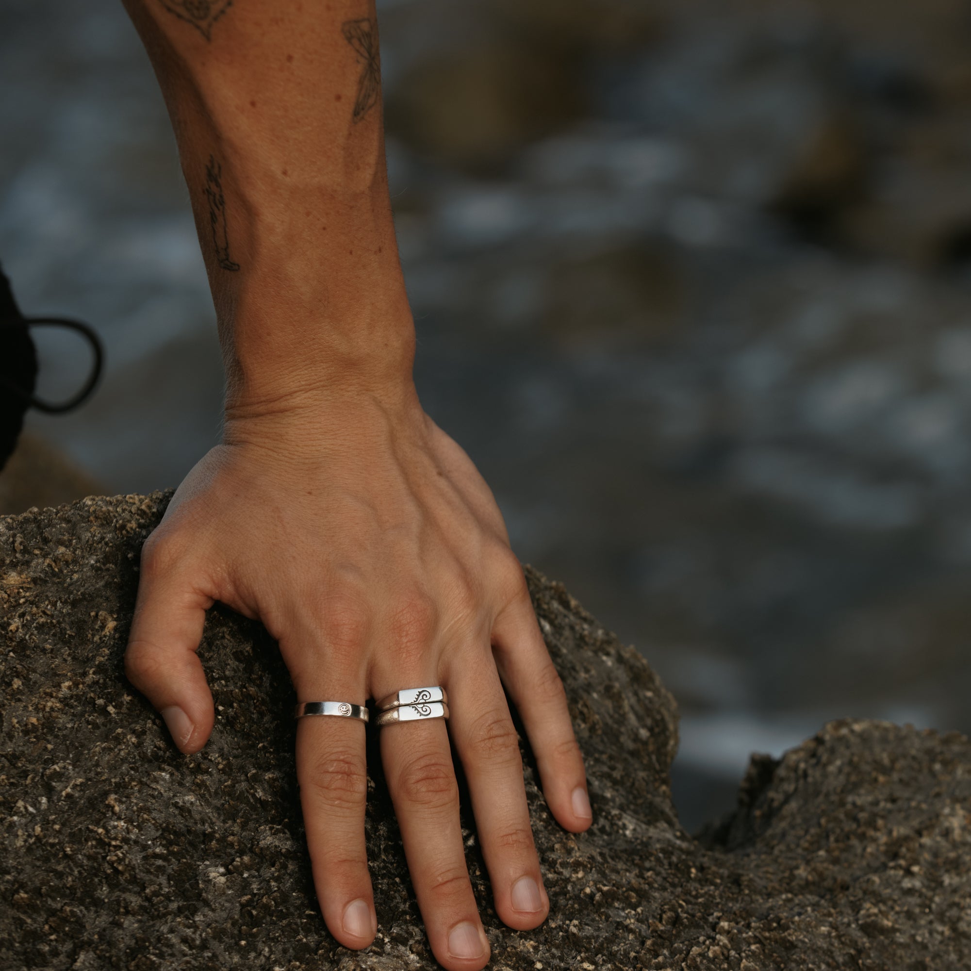 A man's tattooed arm and hand resting on a rock, wearing the Billie Jo Connection Stackers. Men's jewellery for a rugged, modern aesthetic.