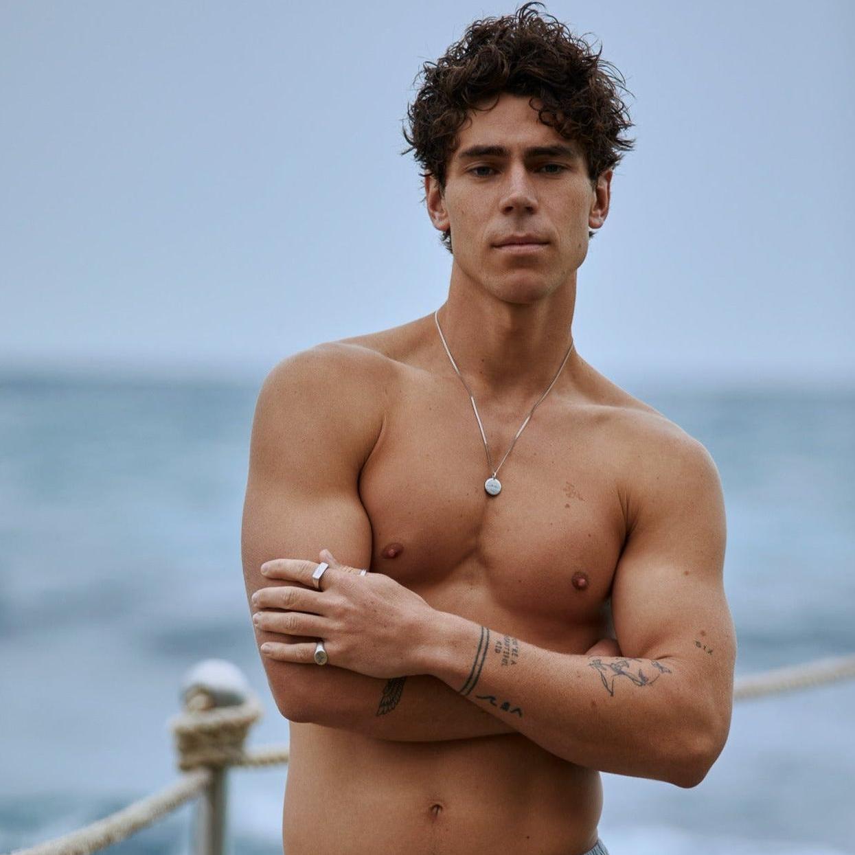 A man models the Billie Jo Untold Pendant on a beach, showcasing the sterling silver necklace against his chest for a modern, masculine look.