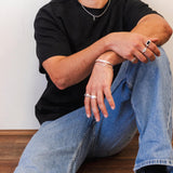 A male model wearing the Billie Jo Canvas Ring and other silver jewellery. He is sitting on the floor, styled in a black t-shirt and blue jeans.
