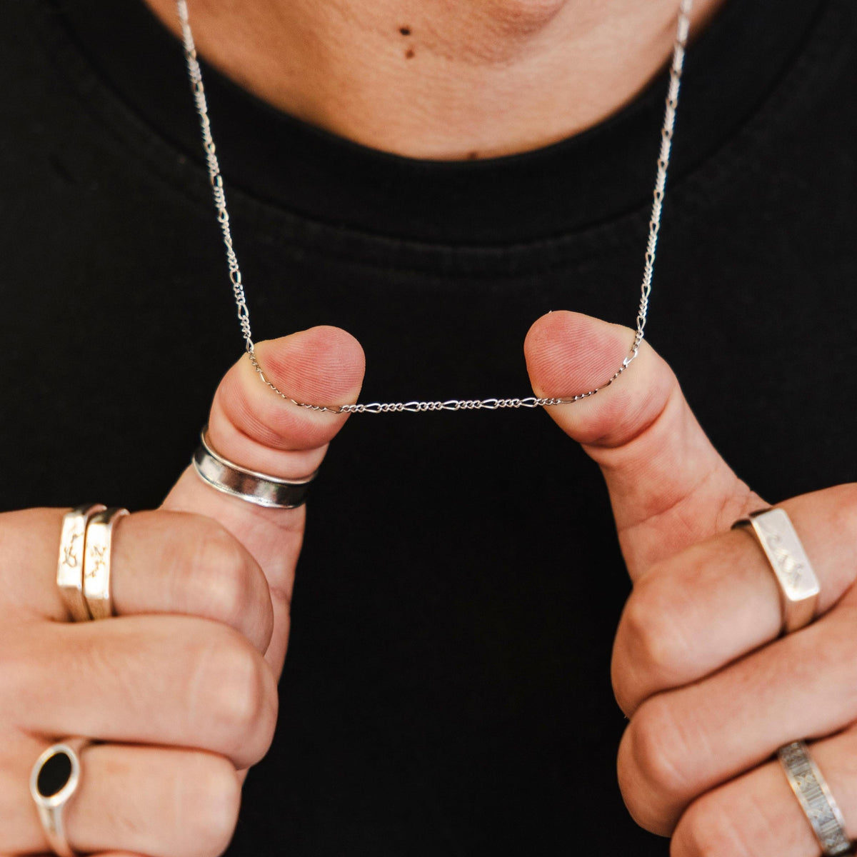 A man's hands, adorned with silver rings, holding up the delicate sterling silver Slim Figaro Necklace by Billie Jo, showcasing the chain's texture and shine.