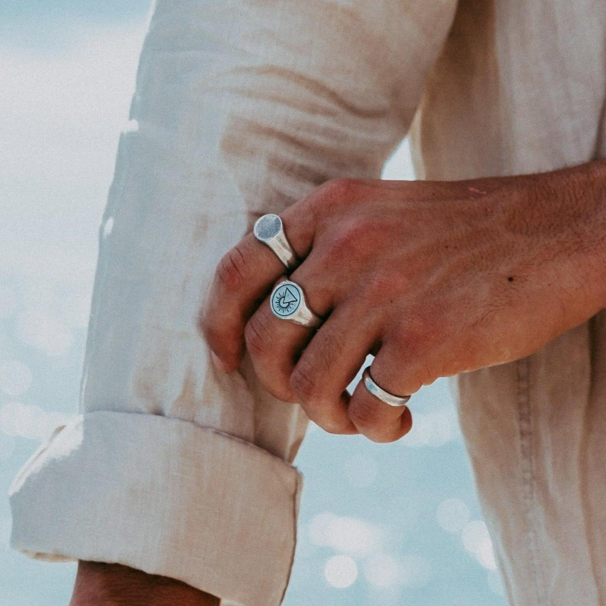 A close-up on a man's hand wearing the Billie Jo Signet Ring and another engraved silver ring, with a blurred background of the ocean.