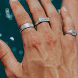 A close-up of a man's hand wearing the Billie Jo Canvas Ring and other sterling silver rings against a sparkling blue water background. A summer lifestyle shot.