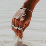 A man wearing the Billie Jo Rope Bracelet in sterling silver, his hand scooping sand on a sunny Gold Coast beach, styled with several silver rings.