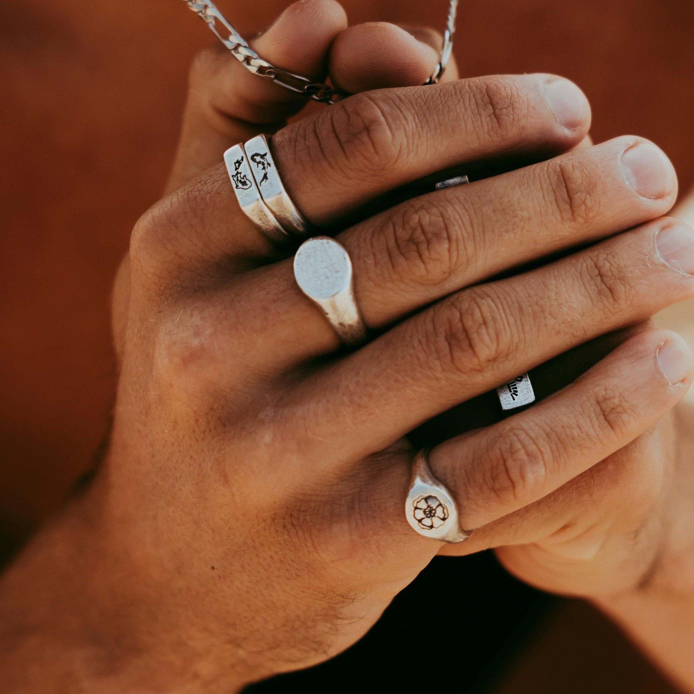 A detailed shot of a man's hands, showing the Billie Jo Signet Ring on his middle finger, stacked with other silver rings and a chain necklace.