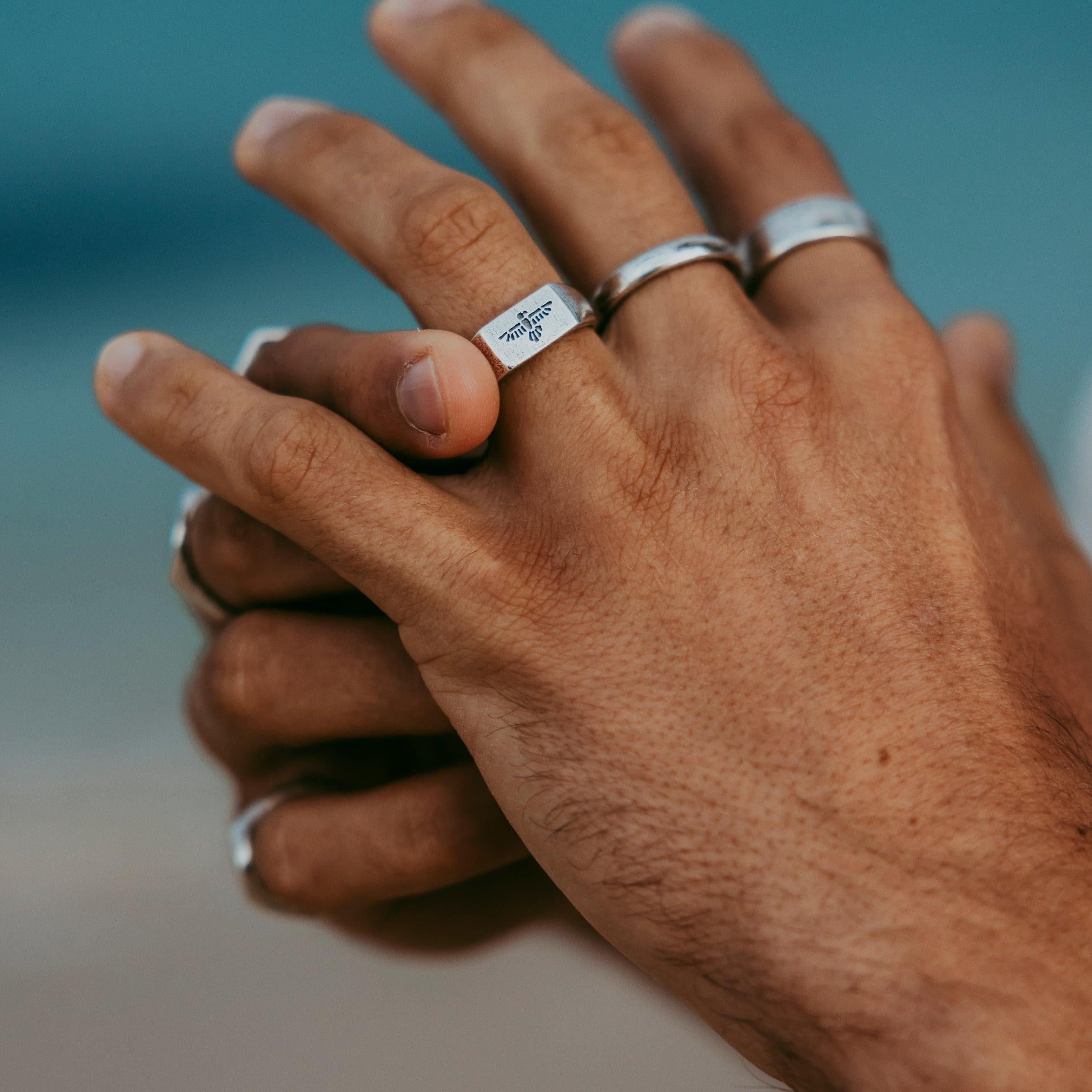 Close-up of the Billie Jo Essence Ring on a man's hand, showcasing the detailed engraving, with a soft-focus beach scene in the background.