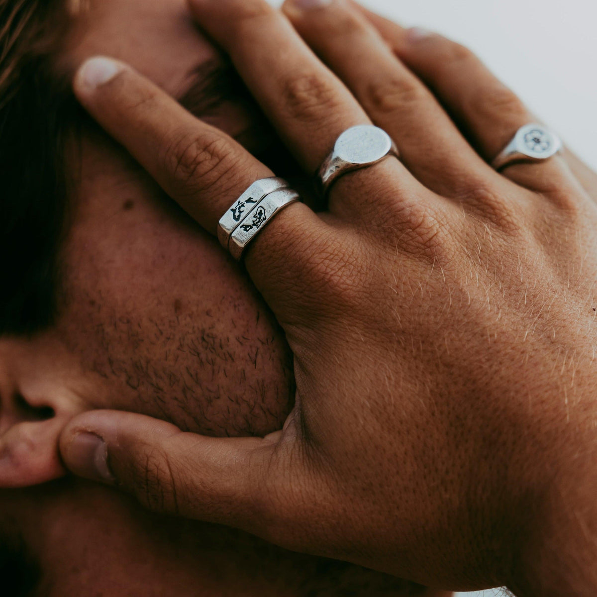 A man's hand covering his face, showing off the Billie Jo KOI I Ring and other silver rings, in a moody, close-up lifestyle shot.