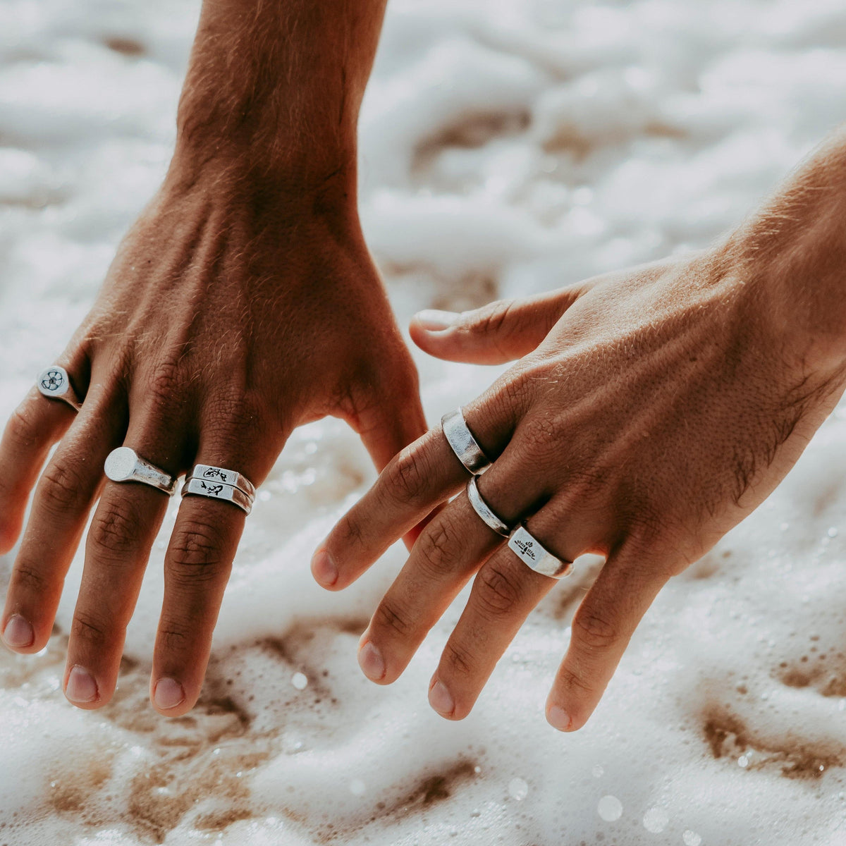 A man's hands in the ocean's white wash, wearing a variety of sterling silver rings from Australian jewellery brand Billie Jo, including the KOI I Ring.