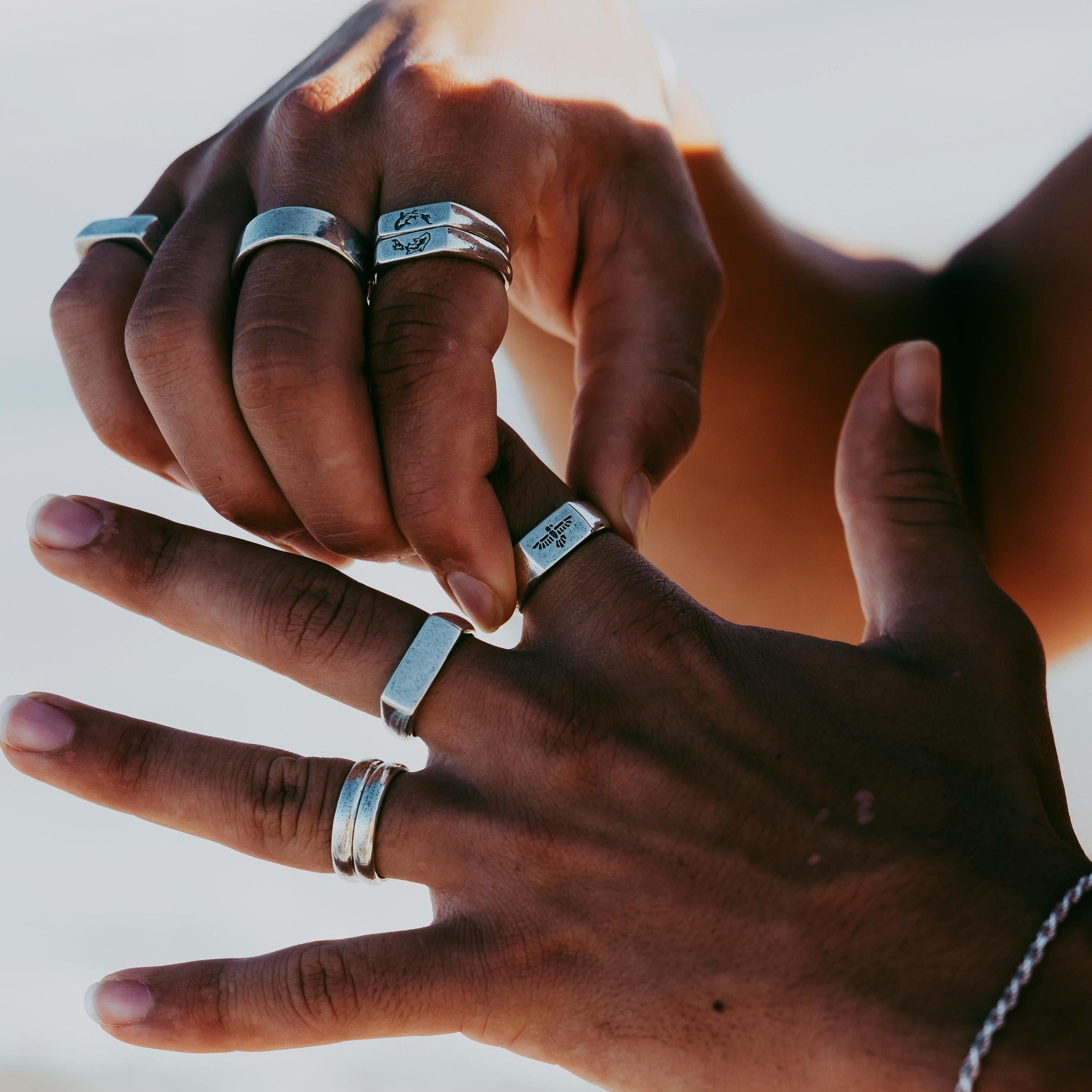 A detailed shot of two hands layered with various silver rings, prominently featuring the engraved Essence Ring by Billie Jo in a bright, sunlit setting.