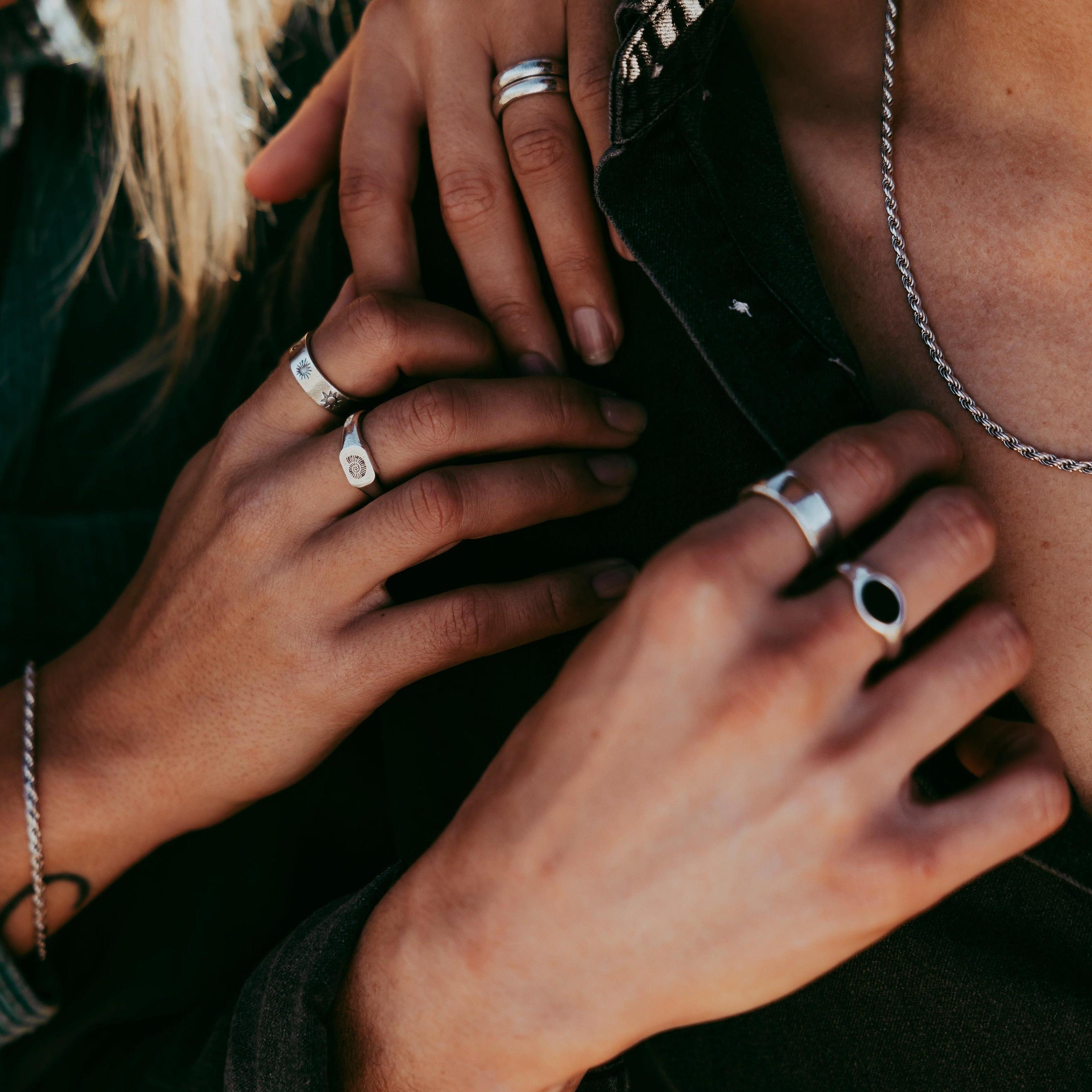 A man wears the sterling silver Billie Jo ONYX 2.0 RING in a close-up shot. Another hand with rings rests on his shoulder, highlighting the jewellery.