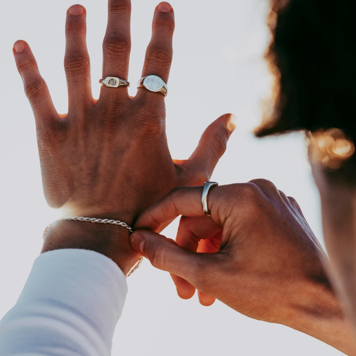 A close-up of a man's hands as he fastens a silver rope bracelet, showing the Billie Jo Signet Ring and another silver ring against a sunlit background.