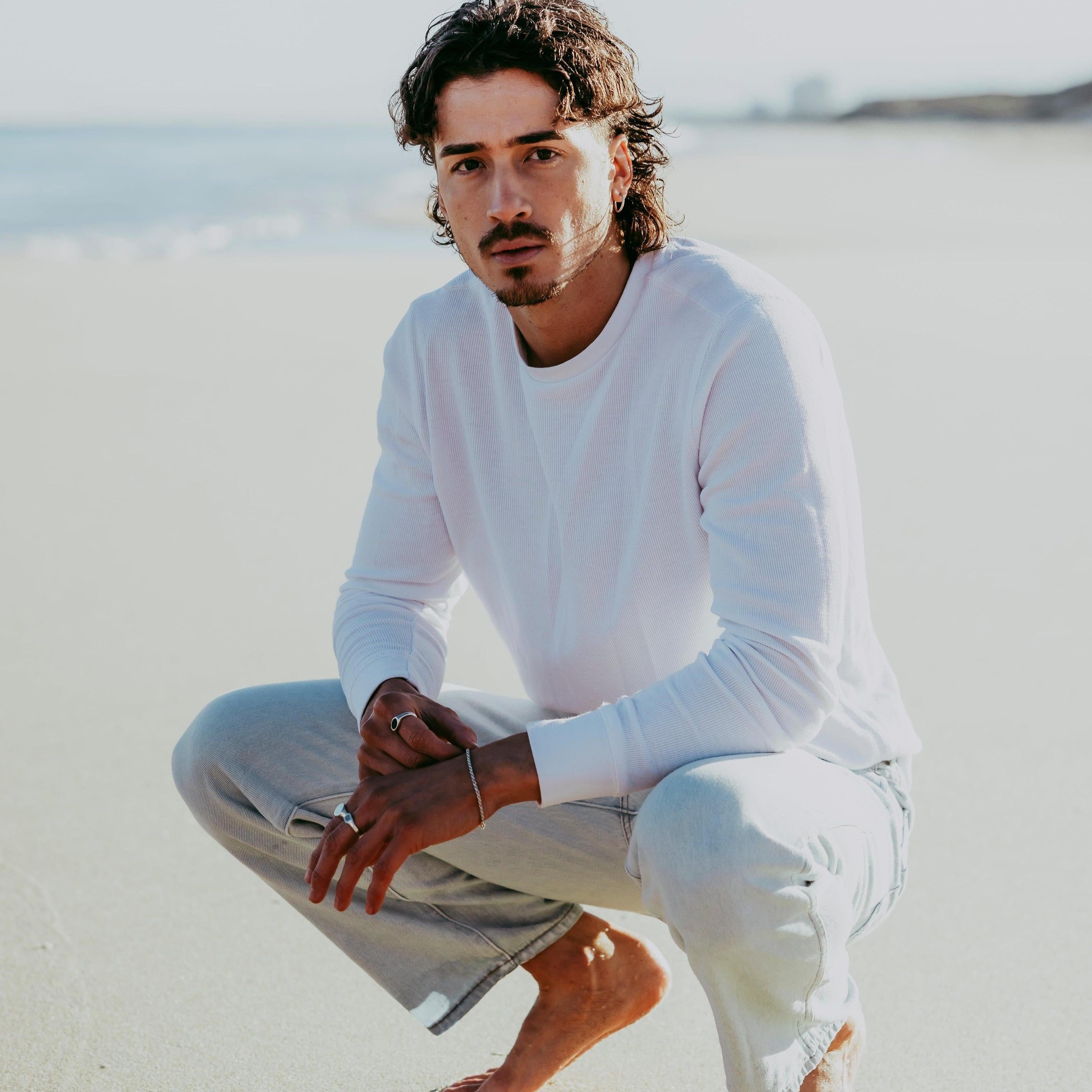 A male model with a mullet hairstyle, wearing the Billie Jo Rope Bracelet and rings, crouching on a sandy beach in a white long-sleeve shirt and light-wash jeans.
