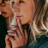 A woman with blonde hair wears the Billie Jo ONYX 2.0 RING, her hand resting on her chin. A man is blurred in the background, creating a romantic, editorial feel for this men's jewellery piece.