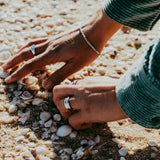 A person wearing a Billie Jo Rope Bracelet sifts through seashells on a sunny beach, the sterling silver of the bracelet glinting in the light.