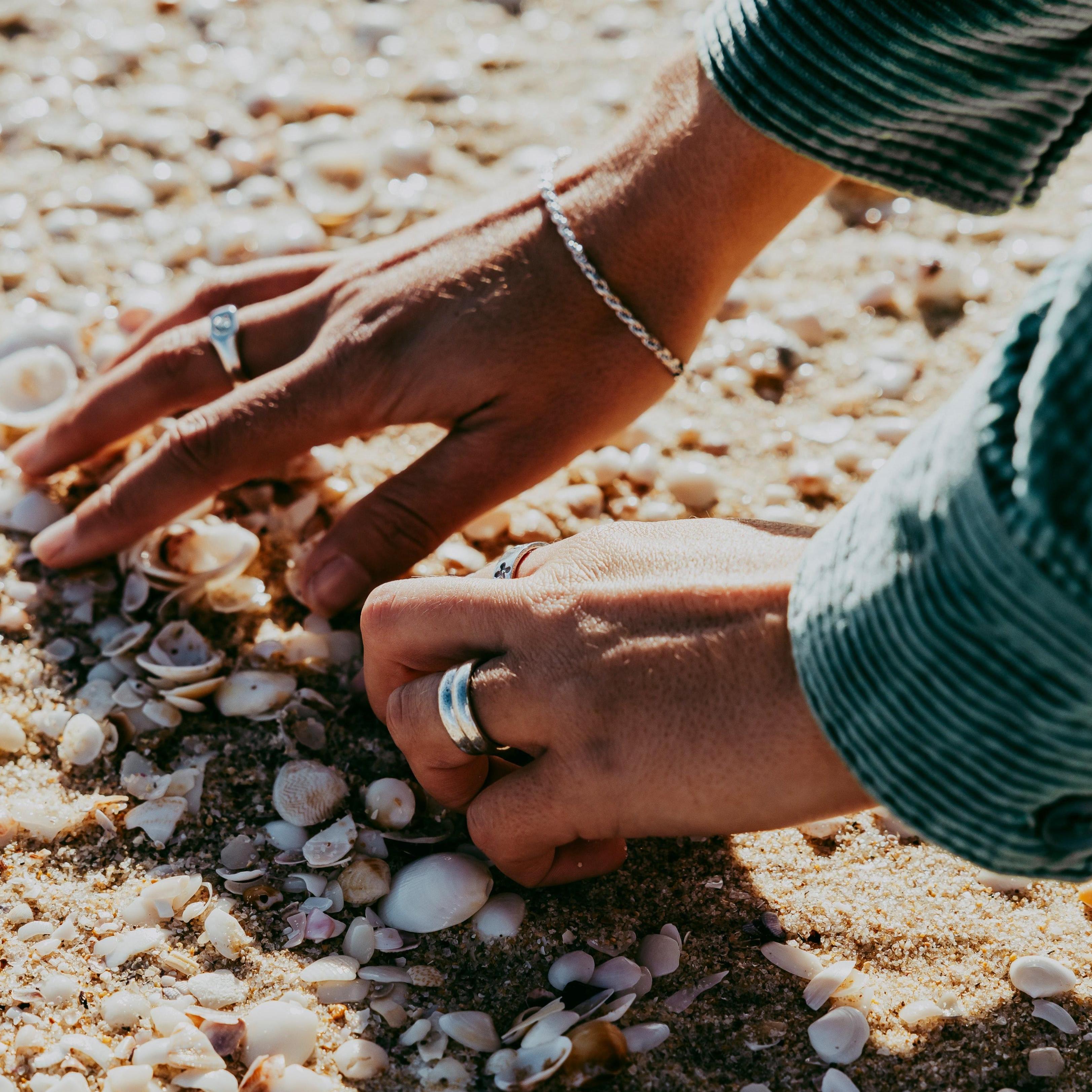 A person wearing a Billie Jo Rope Bracelet sifts through seashells on a sunny beach, the sterling silver of the bracelet glinting in the light.