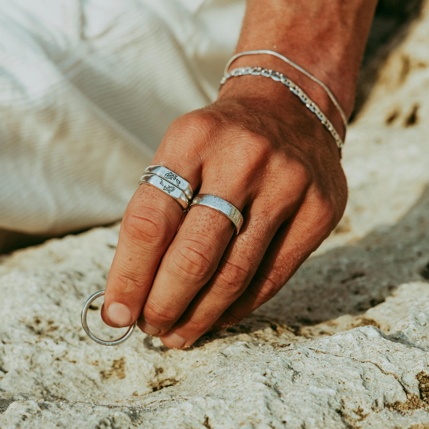 A close-up of a man's hand wearing the Serenity Ring and other silver jewellery from Billie Jo, holding another ring against a rocky, natural backdrop.