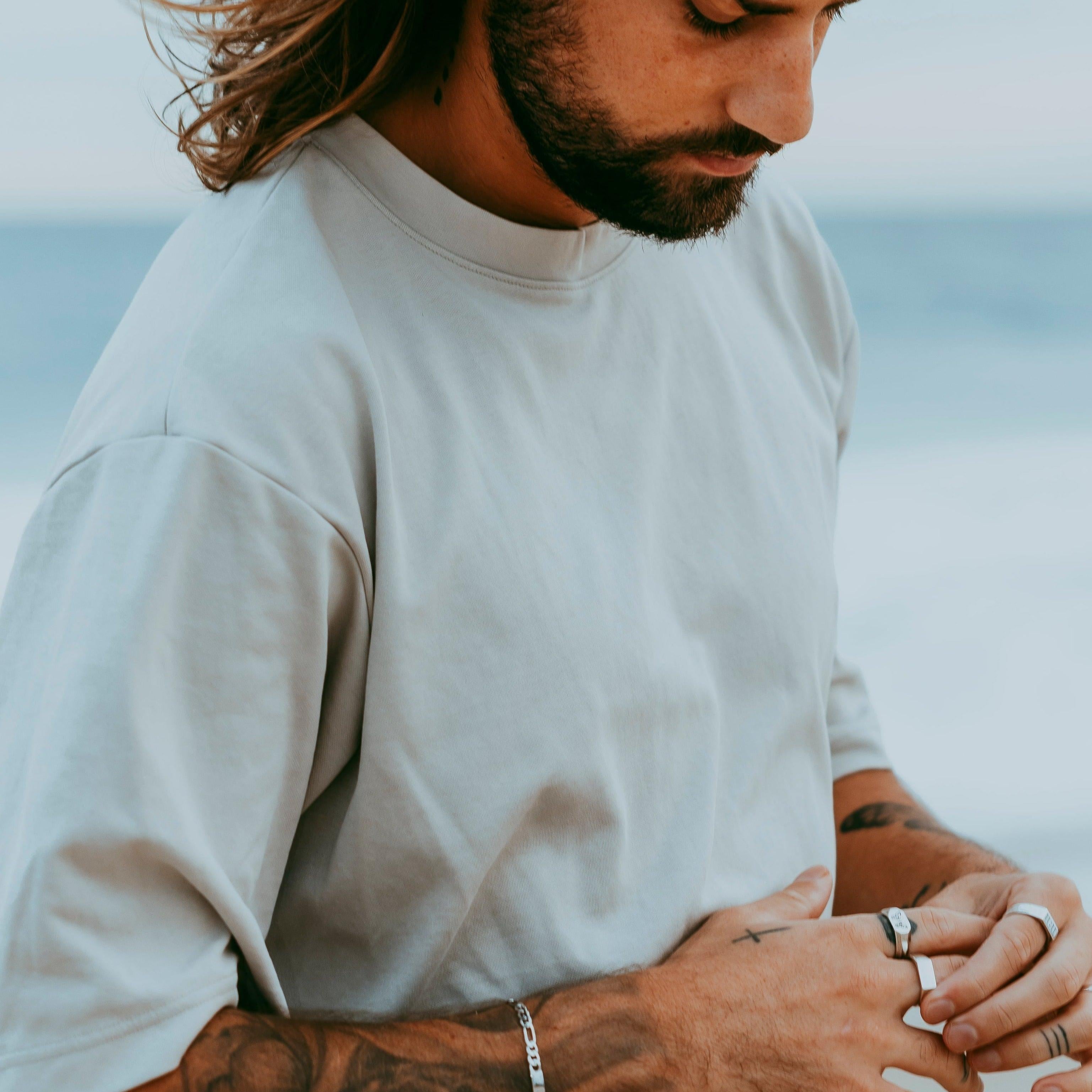 A man wearing the silver Persona Bracelet by Billie Jo, looking down at his hands which are adorned with multiple silver rings, against a beach backdrop.