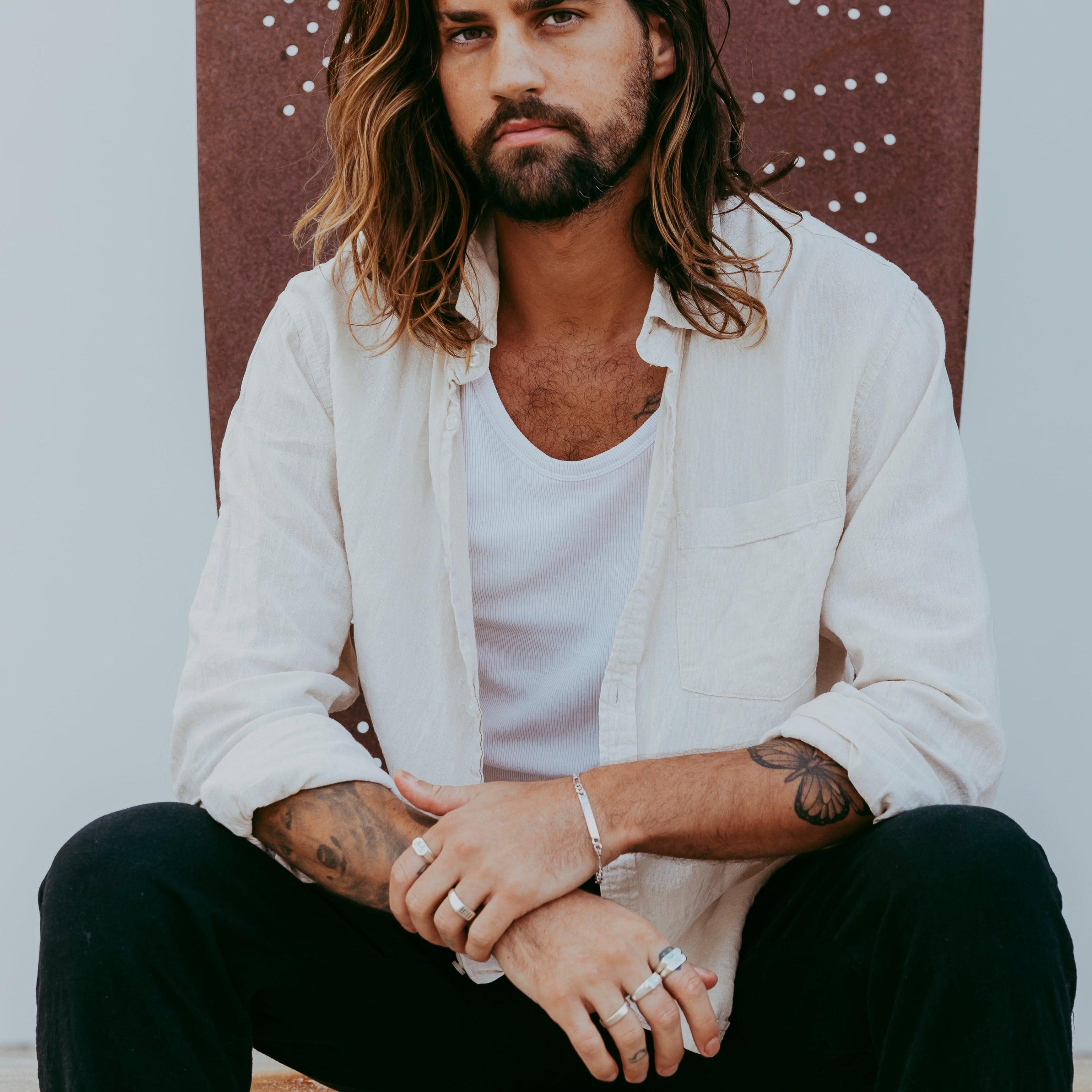 A male model sits wearing the sterling silver Persona Bracelet by Billie Jo, paired with multiple rings and a casual, layered outfit for a modern look.