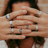 A man with long hair and tattooed fingers holds his hands in front of his face, showcasing a collection of sterling silver rings from Billie Jo, including the Prowess Ring.