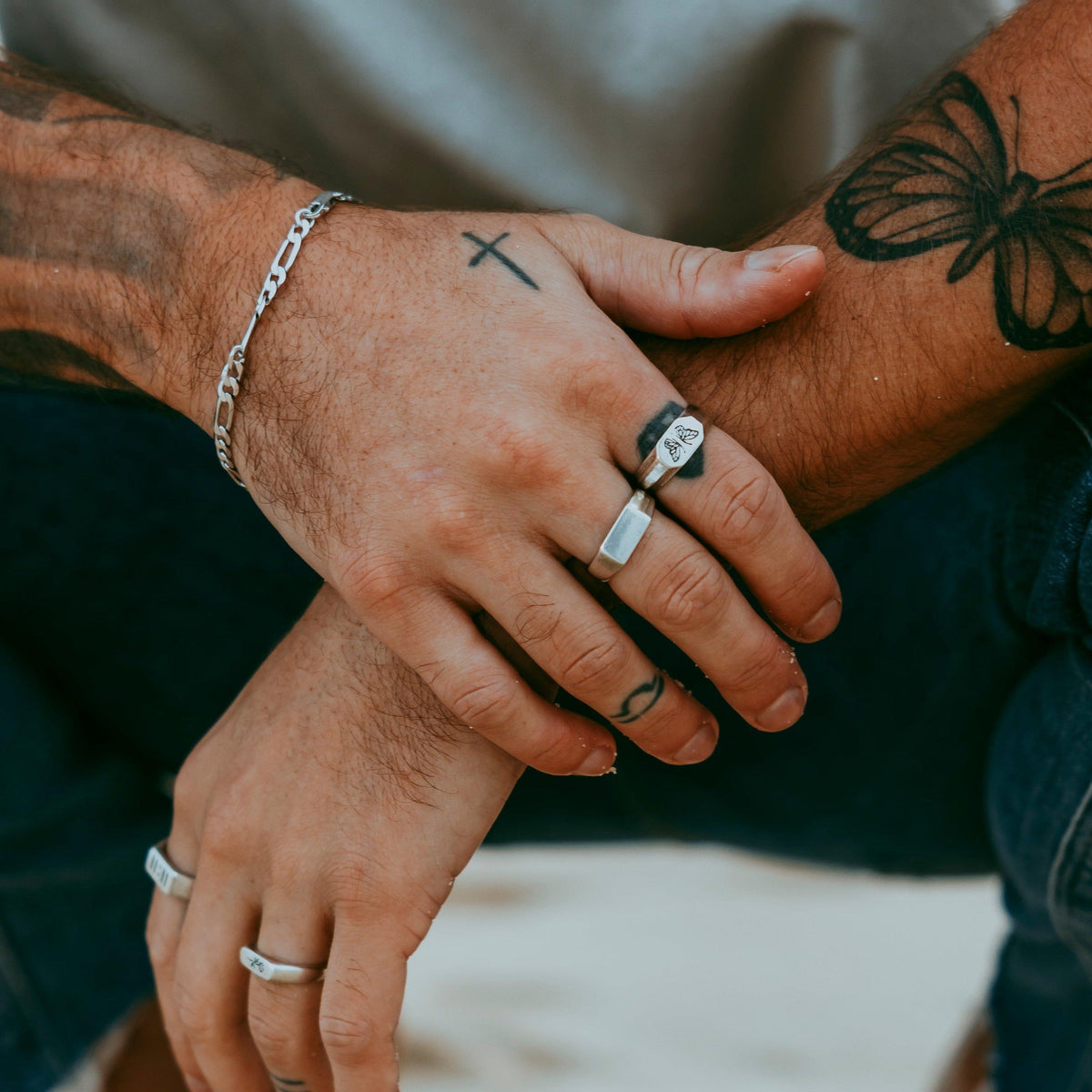 A close-up of a man's tattooed hands, adorned with a stack of sterling silver rings from Billie Jo, including the Genesis Ring. A perfect example of modern men's jewellery styling.