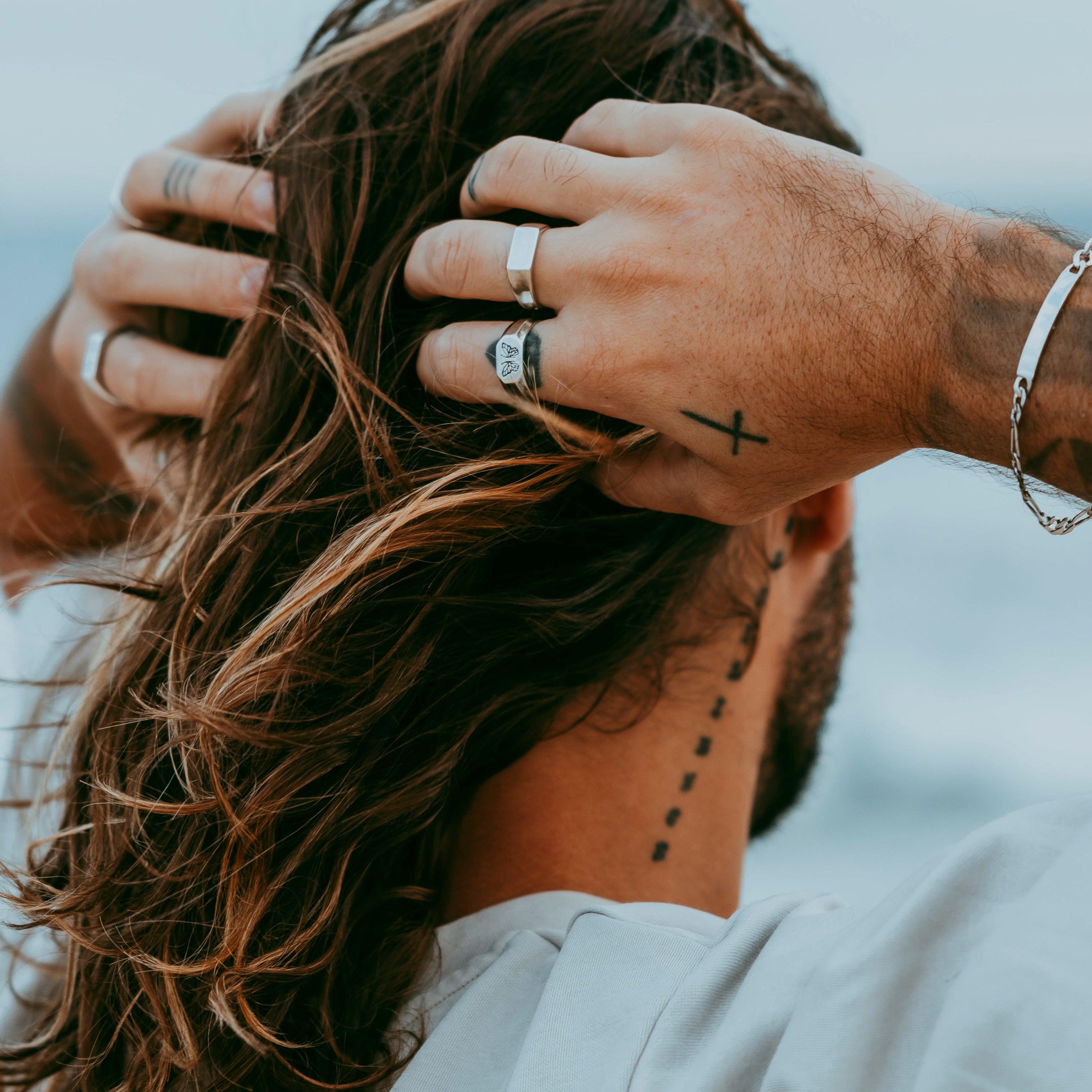 A close-up of a man's hands with tattoos, wearing the Billie Jo Persona Bracelet and several silver rings, running his hands through his hair.