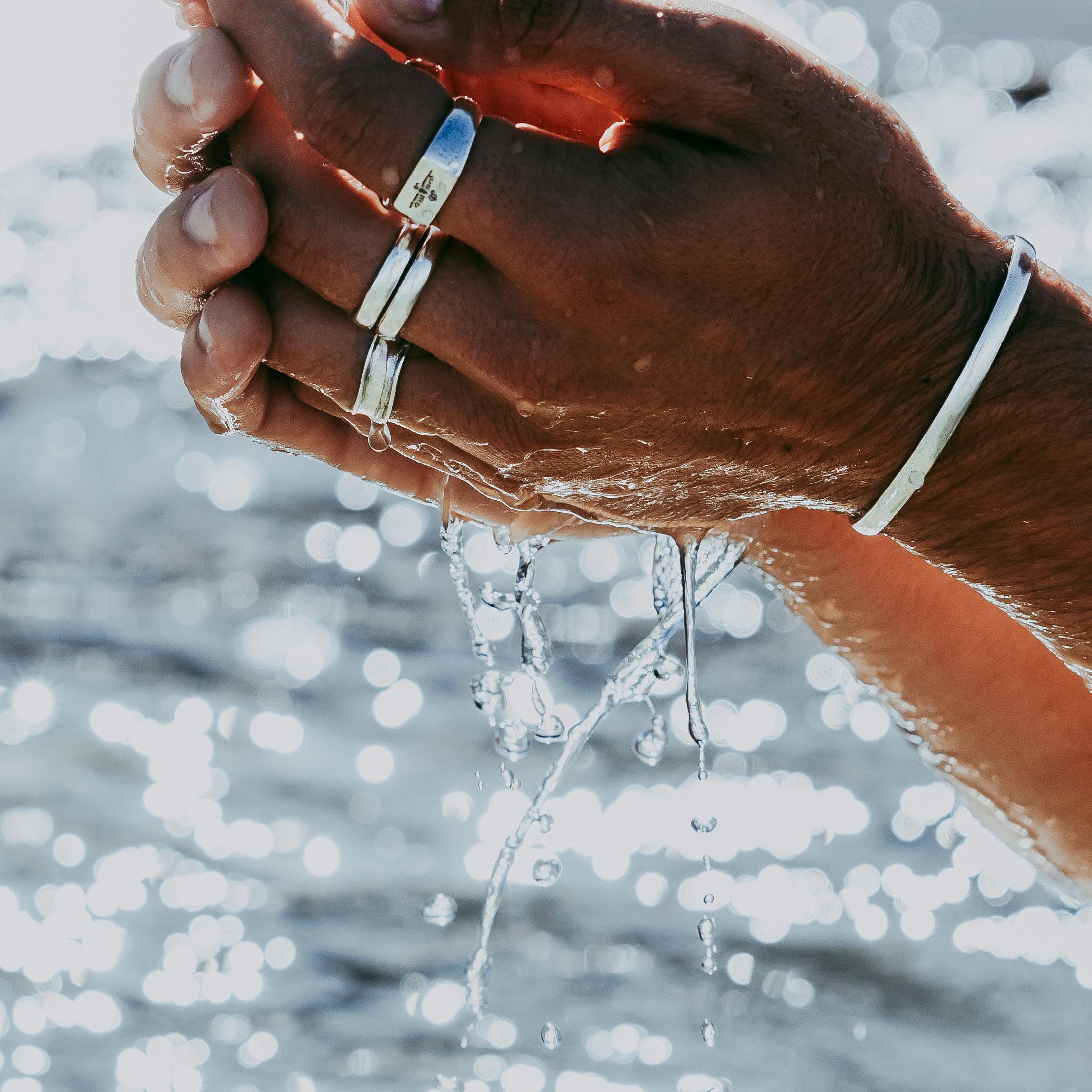 A man's hands, adorned with the silver Essence Ring and a cuff from Billie Jo, cupping water that cascades down against a sparkling ocean backdrop.