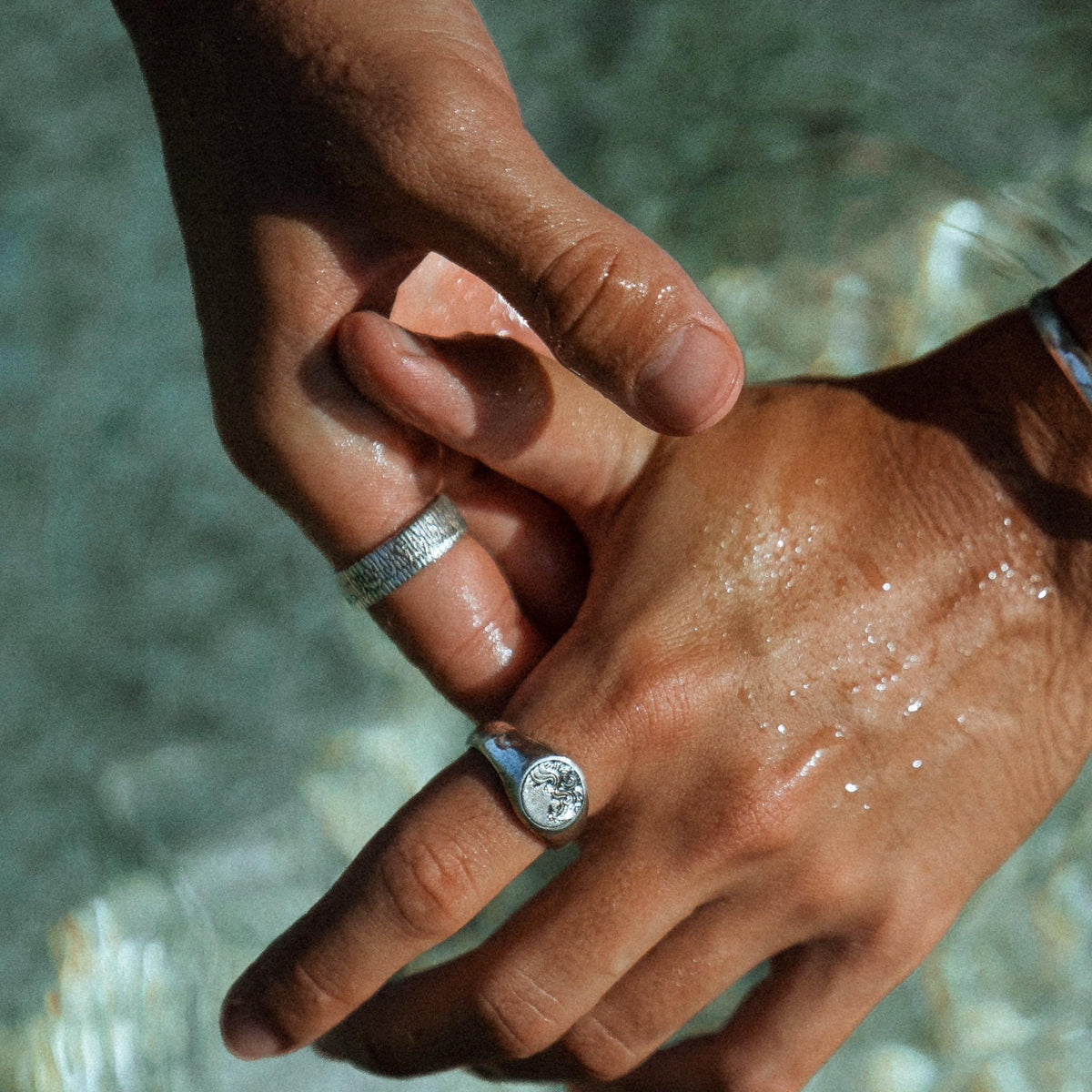 Two hands clasped in water, one wearing the sterling silver Current Ring by Billie Jo, with water droplets glistening on the skin.