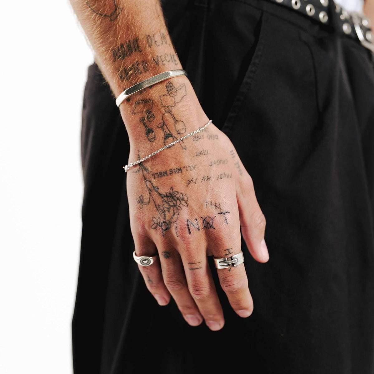 A tattooed man's hand wearing the sterling silver Bind Ring from Billie Jo. He is also wearing a silver cuff and chain bracelet, showcasing a modern, layered jewellery style.