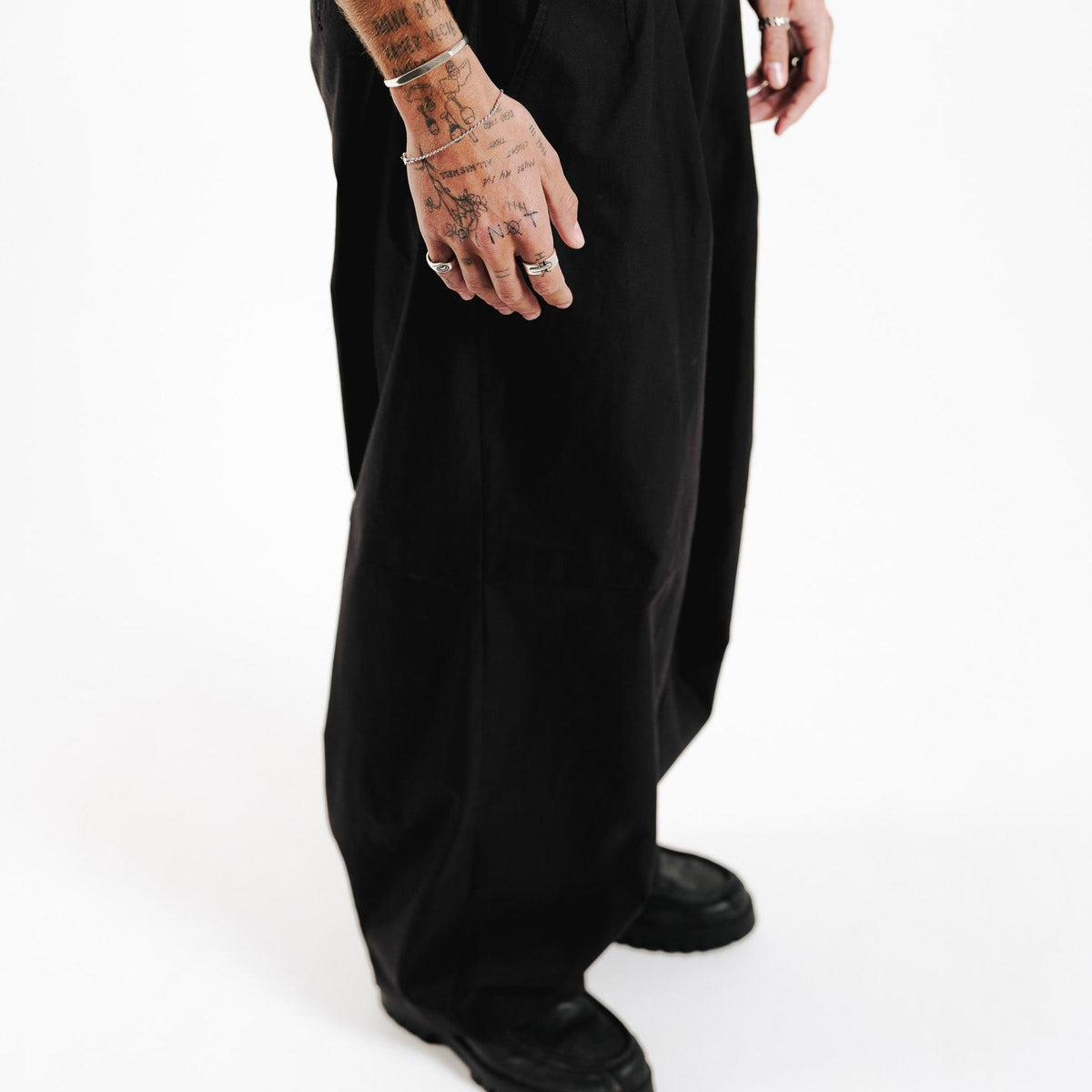 A full-length shot of a man with tattooed hands wearing the Billie Jo Bind Ring. He is dressed in black wide-leg trousers and a black top, showcasing a modern, minimalist style.