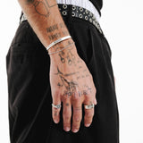 A stylish shot of a man's hand with tattoos, wearing the Billie Jo Slim Rope Bracelet and silver rings, against black trousers for a cool, modern vibe.
