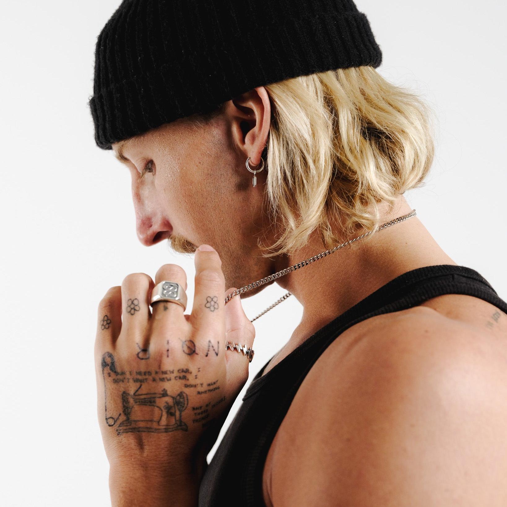 A male model with blonde hair and a black beanie looks down, his face in profile, wearing the Nox Earring from Billie Jo with a silver chain necklace.