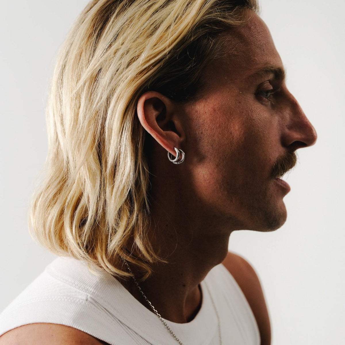 An editorial shot for Billie Jo featuring a man with a moustache and long blonde hair, wearing the Slim Batch sterling silver necklace and silver hoop earrings.
