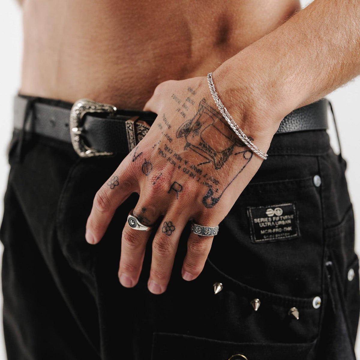 A man with a bare torso and tattooed hands wears the Vita Ring. This lifestyle image for Australian jewellery brand Billie Jo shows the ring styled with black pants and a decorative belt.