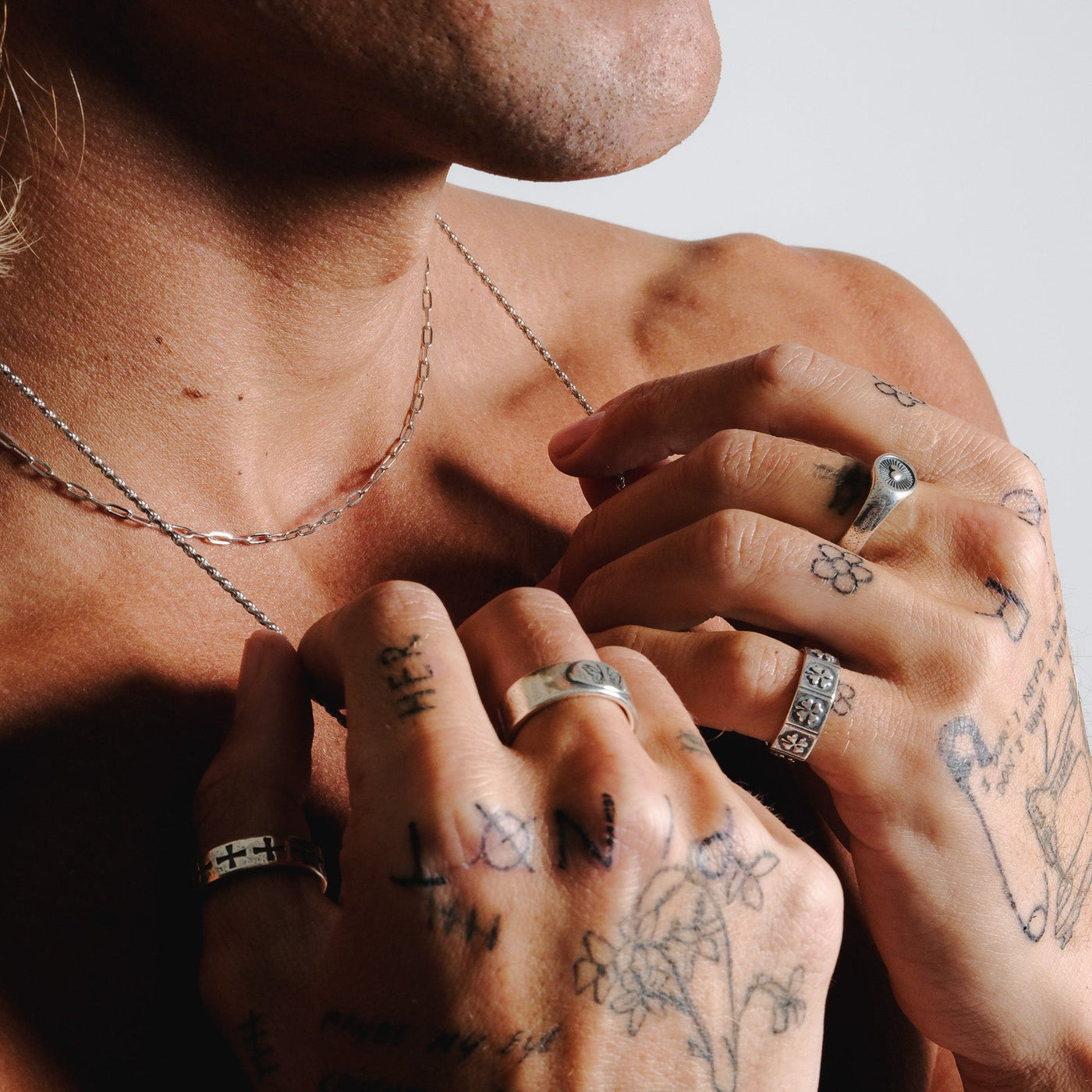 A man with tattooed hands adjusts a silver necklace, wearing the Billie Jo LUCKY RING and other sterling silver rings for a layered jewellery look.