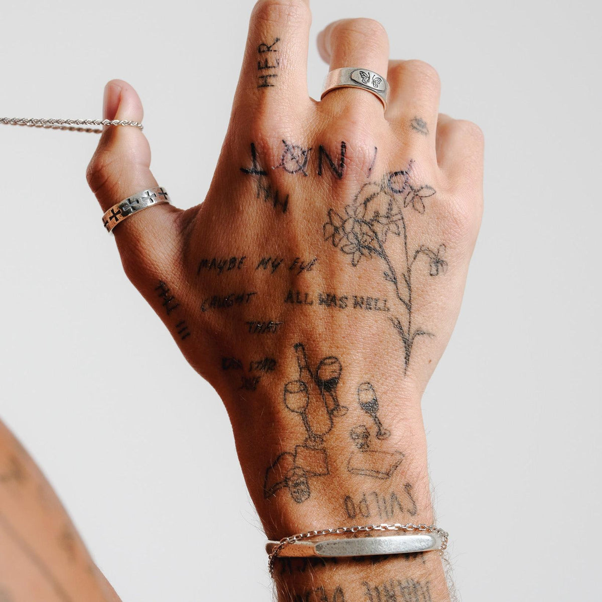 A tattooed hand, adorned with the Billie Jo Sanctum Ring and a silver bracelet, delicately holds a silver chain. An artistic shot showcasing men's jewellery.