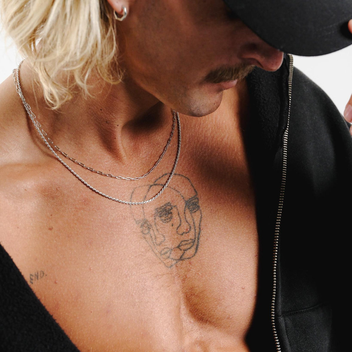 A man with a moustache wears the sterling silver Slim Link Necklace by Billie Jo, layered with a rope chain and styled with a black hoodie and cap.