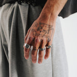 A man's tattooed hand wearing the sterling silver Vita Ring from Australian jewellery brand Billie Jo. The ring is styled with other silver rings against a background of grey pants, showcasing its versatility.