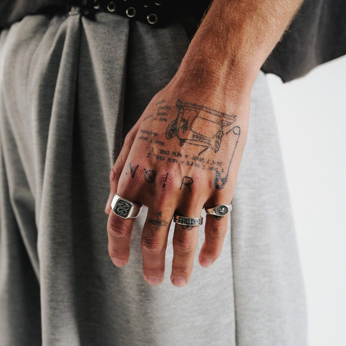 A man's tattooed hand, wearing the Billie Jo Serpent Ring and two other silver rings, against a backdrop of grey trousers and a black t-shirt.