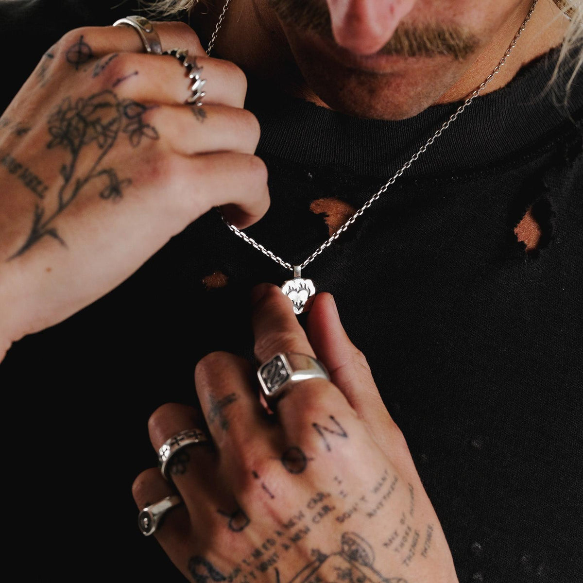 A man with tattooed hands adjusts the Ember Pendant, providing a close-up of the sterling silver piece against his black shirt.