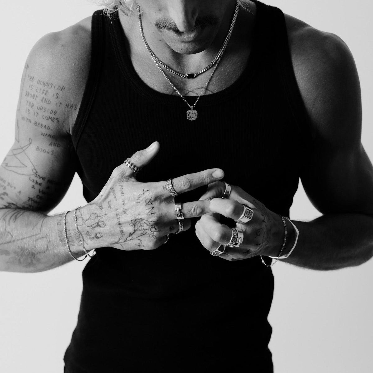 A black and white editorial shot of a man with tattooed hands wearing multiple silver rings, including the Billie Jo Bind Ring. He is wearing a black singlet and has several necklaces on.