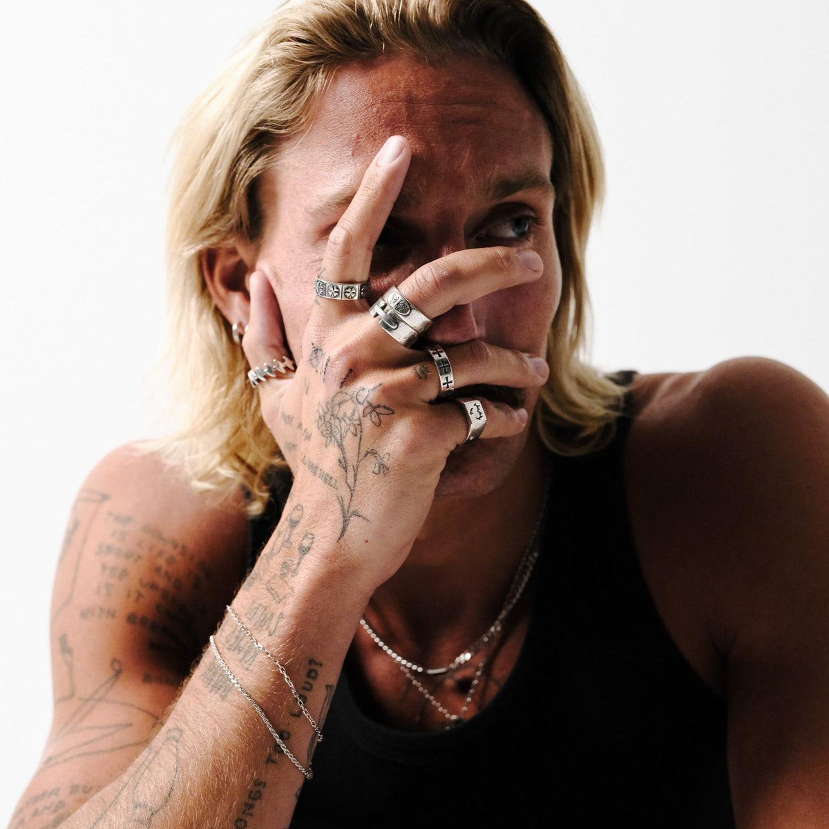 A male model with blonde hair and tattoos showcases the Billie Jo LUCKY RING as part of a stacked silver ring look, embodying the brand's modern and edgy aesthetic.