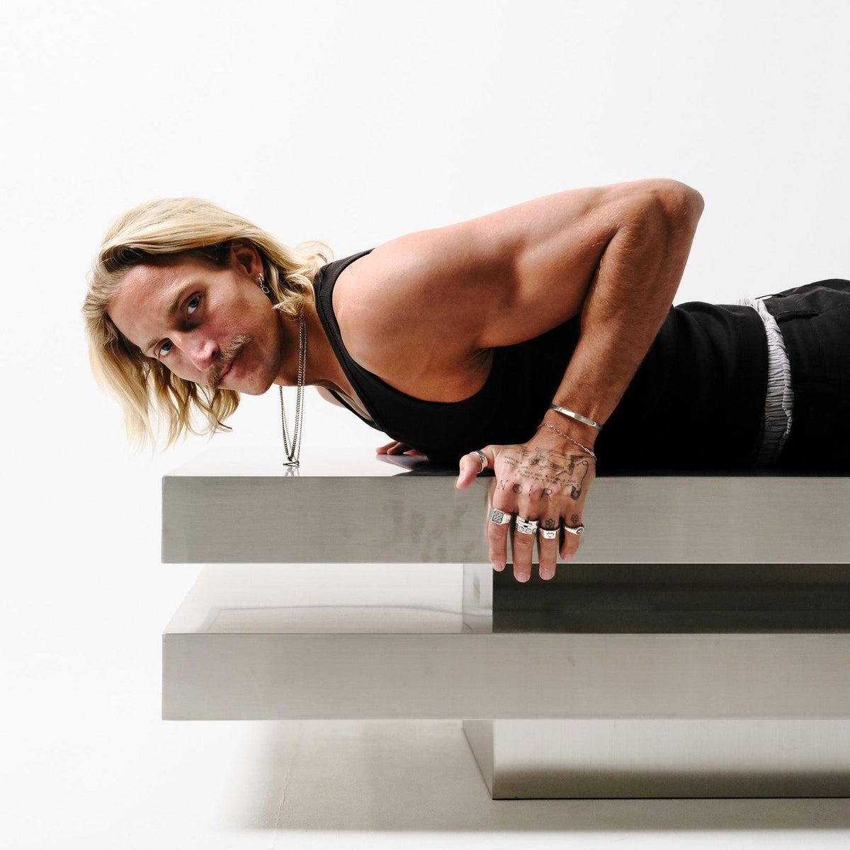 A male model with long blonde hair and a moustache, wearing the Billie Jo Echo Cuff, a necklace, and multiple rings, leans over a modern, tiered table. An editorial shot for this Australian men's jewellery brand.