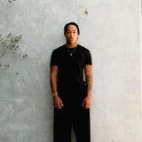 Full-length shot of a male model wearing the Billie Jo Paradiso Pendant with a black t-shirt and trousers against a concrete wall. Men’s jewellery.