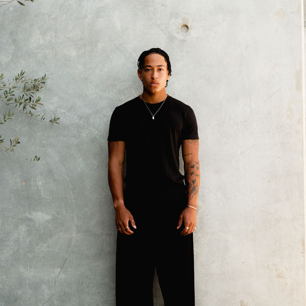 Full-length shot of a male model wearing the Billie Jo Paradiso Pendant with a black t-shirt and trousers against a concrete wall. Men’s jewellery.