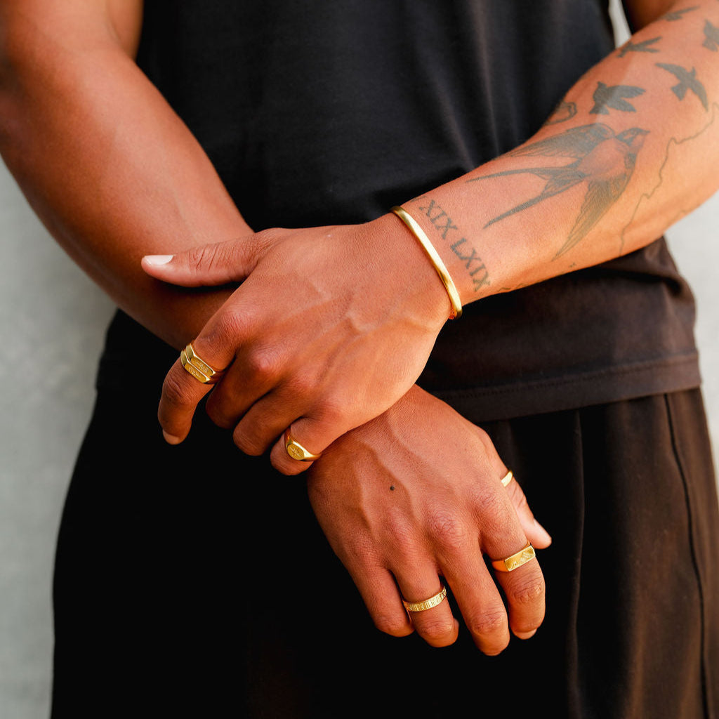 A detailed shot of the Billie Jo Drifter Cuff in gold on a man's tattooed arm, paired with a stack of gold rings for a bold statement.
