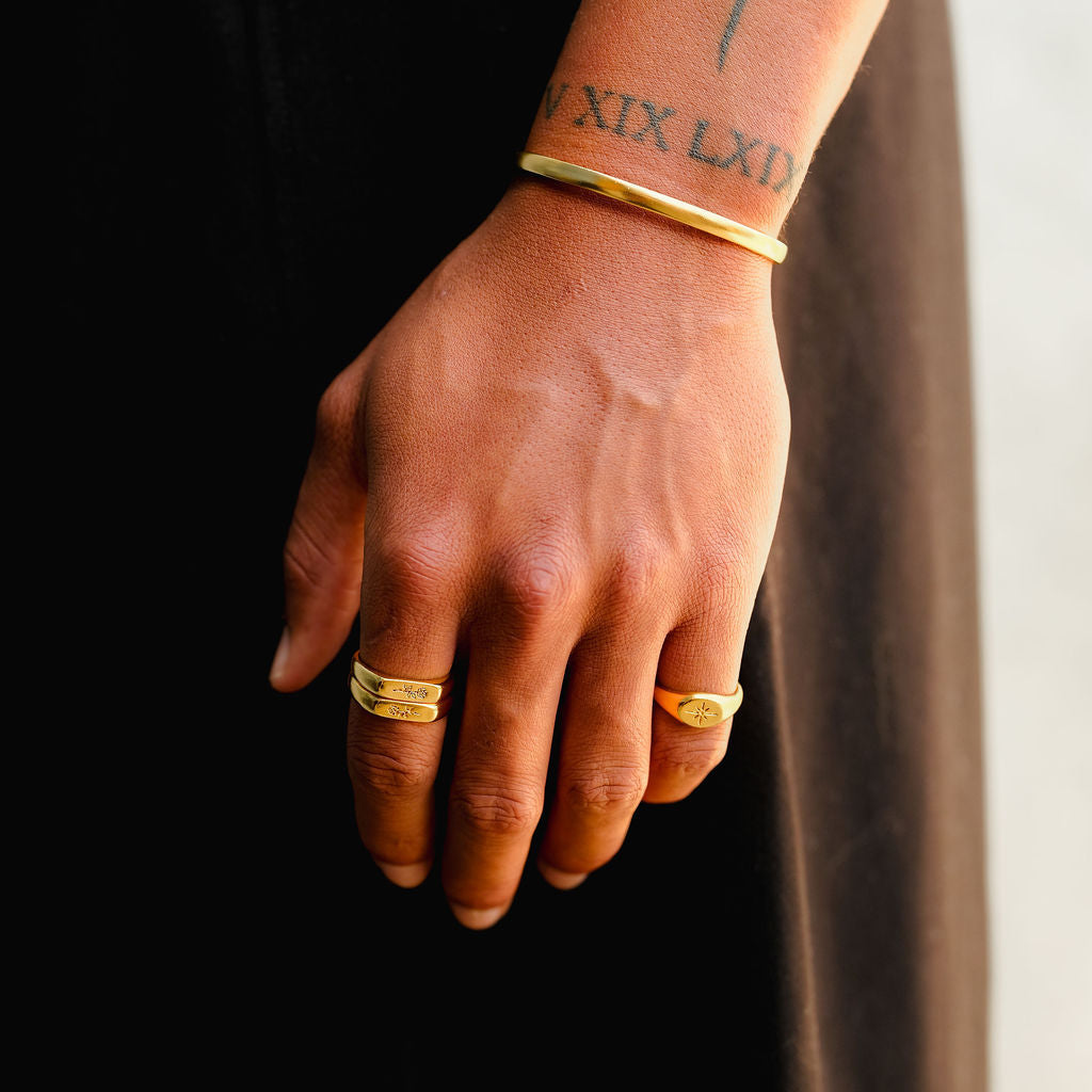A man's tattooed arm and hand, wearing the gold Rosa Stacker rings and a gold cuff from Billie Jo, against a dark, modern backdrop.