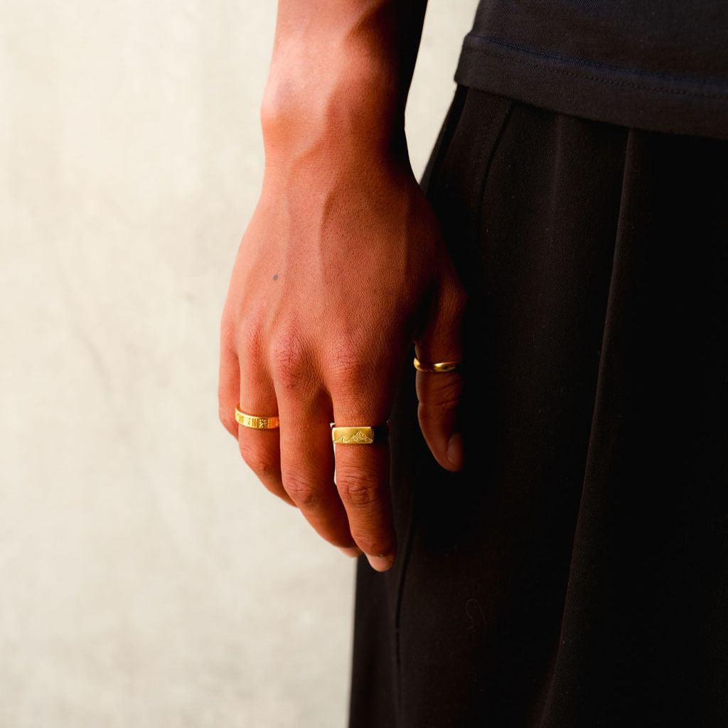 A man's hand, adorned with the Gold Relic Ring and other gold jewellery from Billie Jo, resting against a black fabric background. A minimalist and modern style.