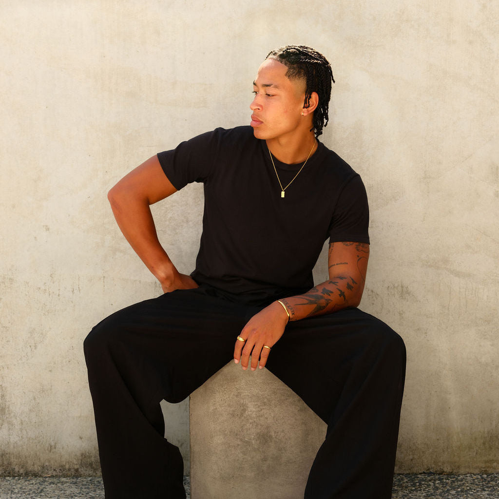 A male model wears the gold Paradiso Pendant from Billie Jo, styled with a black t-shirt and trousers for a modern, minimalist look. Gold Coast jewellery.