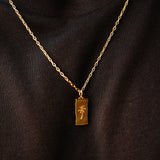 Close-up of the Billie Jo Paradiso Pendant in gold, showing the detailed palm tree engraving on the rectangular tag against a black fabric background.