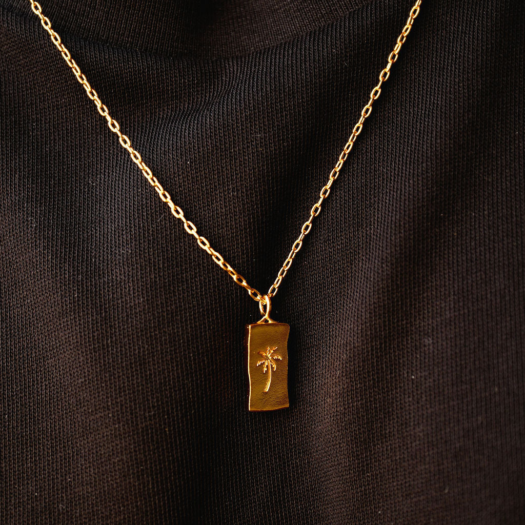Close-up of the Billie Jo Paradiso Pendant in gold, showing the detailed palm tree engraving on the rectangular tag against a black fabric background.