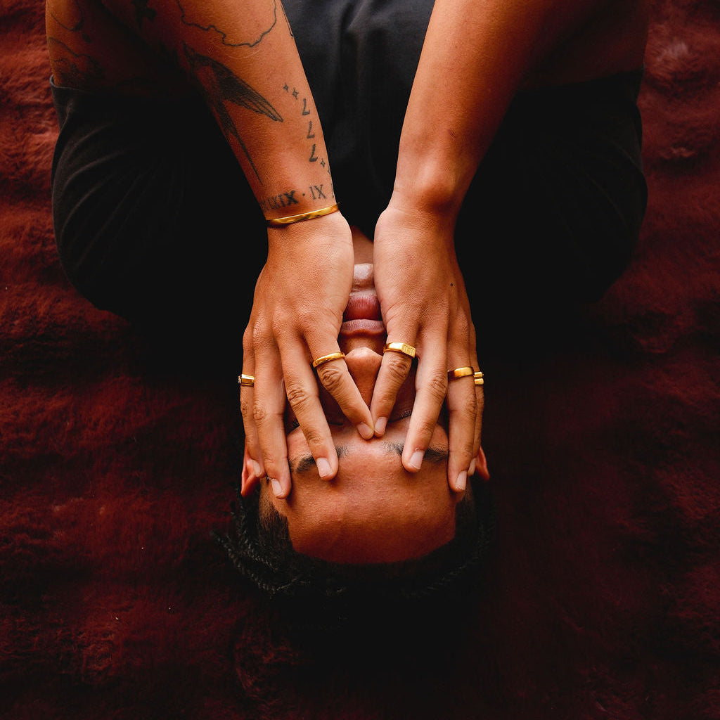 A man with tattooed arms covers his face with his hands, which are adorned with a variety of gold rings from Billie Jo, including the Stacker Ring. An atmospheric, editorial shot.